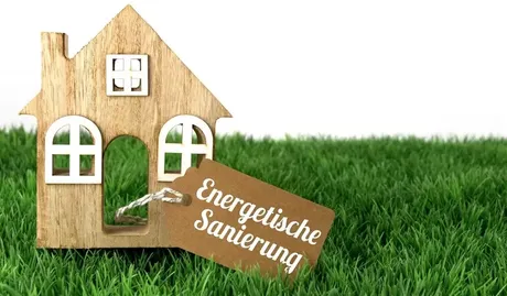 Grüne Wiese mit einem kleinen Spielzeug Holzhaus und einem Etikett mit der Beschriftung Energetische Sanierung.
