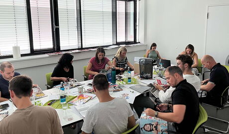 Menschen sitzen um einen Tisch und schneiden aus Zeitschriften Artikel aus.